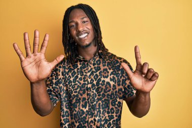 Saçları örgülü, leopar desenli hayvan desenli tişörtlü Afro-Amerikalı adam yedinci parmaklarını gösterip gösterirken kendinden emin ve mutlu gülümsüyor.. 