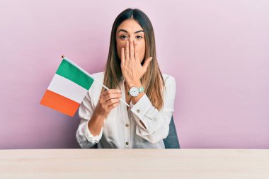 Genç İspanyol kadın İrlanda bayrağını elinde tutarak masanın üzerinde oturmuş, ağzı eliyle kapatıyordu, şok olmuştu ve hata yapmaktan korkuyordu. şaşırmış ifade 
