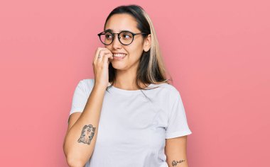 Gündelik beyaz tişört giyen genç İspanyol kadın gergin ve gergin görünüyor. Ağzı tırnak yiyen ellerle. anksiyete. 