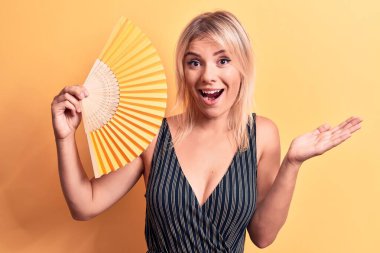 Sarışın, güzel bir kadın el fanı kullanıyor. Sarı arka planda duruyor. Başarımızı kutluyoruz.