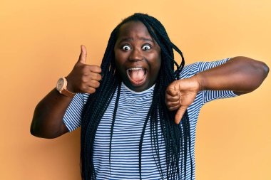 Örgülü genç siyahi kadın başparmağını kaldırıyor ve başparmağını kaldırıyor. Çılgınlığı kutluyor ve başarıyı hayretle kutluyor. Gözleri açık, heyecanla haykırıyor.. 