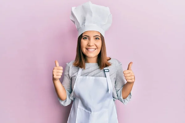 年轻漂亮的女人穿着职业厨师制服 戴着帽子成功的标志 用手做着积极的手势 竖起大拇指 笑容满面 快乐无比 快乐的表达和胜利的姿态 — 图库照片