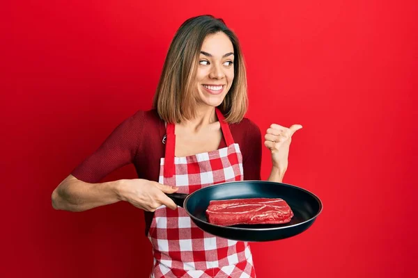 若いですCaucasianブロンド女性身に着けていますクックエプロン調理肉上のパンポインティング親指上のザ サイド笑顔幸せとともにオープン口 — ストック写真