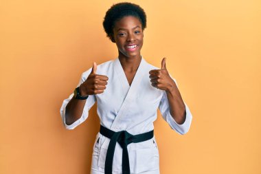 Genç Afrikalı Amerikalı kız karate kimonosu ve siyah kuşak başarısı işareti takıyor. Elleriyle olumlu jest yapıyor. Gülümsüyor ve mutlu. Neşeli ifade ve kazanan jest. 