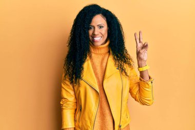 Orta yaşlı Afro-Amerikalı kadın yün kışlık kazak ve deri ceket giyer ve parmaklarıyla iki numarayı gösterirken kendinden emin ve mutlu bir şekilde gülümser.. 