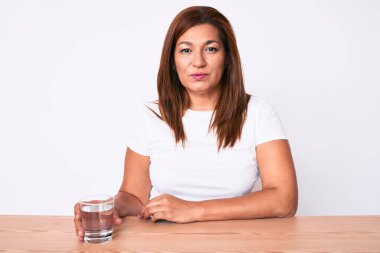 Orta yaşlı esmer kadın masaya oturmuş bir bardak su içiyor düşünceli tavır takınıyor ve kendinden emin görünüyor. 