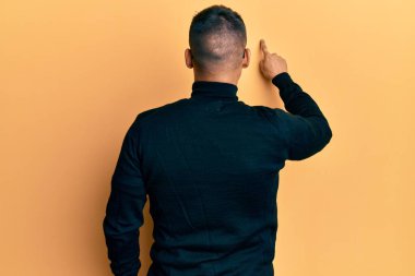 Balıkçı yaka kazak ve gözlük takan yakışıklı bir adam parmak eliyle geriye doğru poz veriyor. 