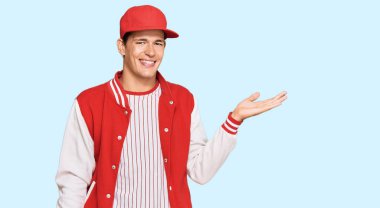 Beyzbol üniforması giyen yakışıklı, beyaz bir adam. Neşeli bir şekilde gülümsüyor ve avuç içi kameraya bakıyor.. 