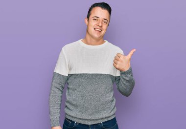 Kış süveteri giyen yakışıklı genç adam gülümseyen yüz ifadesiyle ve baş parmağıyla yan tarafa bakıyor.. 