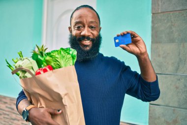 Sakallı Afrikalı Amerikalı bir adam elinde süpermarketten alınmış kağıt torbalarla kredi kartı gösteriyor.