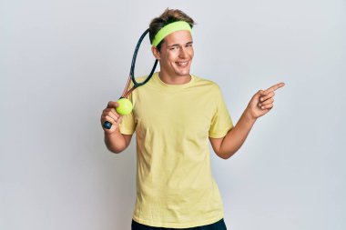 Tenis oynayan yakışıklı beyaz adam raket ve top tutuyor. Gülümsüyor, mutlu bir şekilde elini ve parmağını yana doğru gösteriyor. 