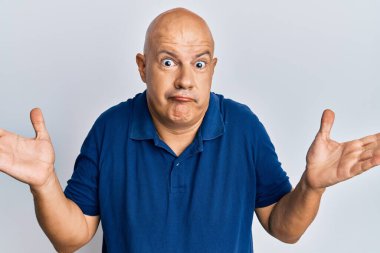 Orta yaşlı, kel, sıradan kıyafetler giyen, kafası karışmış, kolları açık, hiçbir fikri olmayan, şüpheli bir yüzü olan bir adam.. 