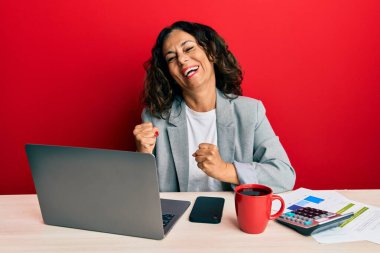 Ofiste çalışan orta yaşlı güzel bir kadın bir fincan kahve içerken çok mutlu ve heyecanlı bir şekilde kollarını kaldırıp, gülümseyerek ve başarı için çığlık atarak kazanan bir jest yapıyor. kutlama konsepti. 