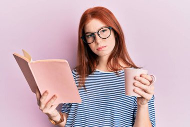 Genç kızıl saçlı kız kitap okuyor ve bir fincan kahve içiyor şüpheci ve gergin, sorun yüzünden surat asıyor. negatif kişi. 