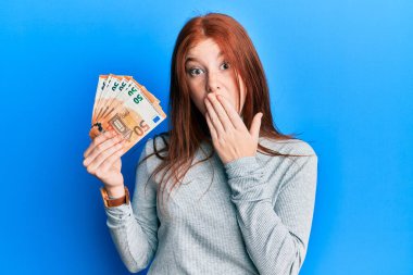 Genç kızıl saçlı kız 50 Euro 'luk banknotları elinde tutuyor. Ağzı ağızla kapatıyor, şok geçiriyor ve hata yapmaktan korkuyor. şaşırmış ifade 