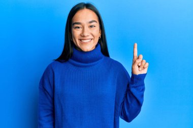 Genç Asyalı kadın, günlük kışlık süveter giyiyor ve bir numaralı parmağını gösterip gülümserken kendinden emin ve mutlu.. 