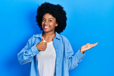 Genç Afro-Amerikalı kadın, günlük giysiler giymiş, avuç içini gösteriyor ve baş parmaklarıyla iyi bir jest yapıyor, mutlu ve neşeli gülümsüyor. 