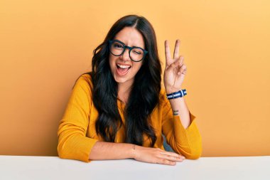 Güzel, esmer, gözlüklü, sıradan giysiler giyen, masaya oturmuş, gülümseyen, kameraya göz kırpan, zafer işareti yapan genç bir kadın. İki numara.. 