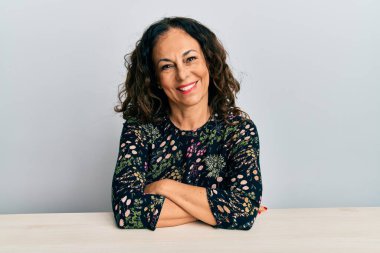 Masada oturan, kollarını kavuşturup kameraya bakan ve gülümseyen orta yaşlı güzel bir kadın. pozitif kişi. 