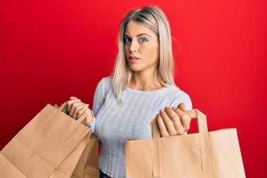 Güzel sarışın kadın karton alışveriş torbalarını şok suratıyla tutuyor, şüpheci ve alaycı görünüyor, ağzı açık kalmış. 