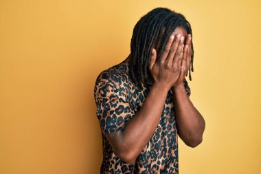 Saçları örgülü, leopar desenli hayvan desenli tişörtlü, suratını ellerle örten ve ağlayan Afro-Amerikan bir adam. depresyon kavramı. 