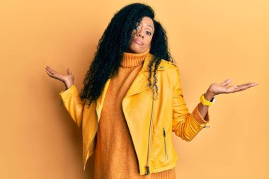 Orta yaşlı Afro-Amerikalı kadın yün kazak ve deri ceket giyiyor ve elleri ve kolları havada, kafası karışmış bir ifade takınıyor. şüphe kavramı. 