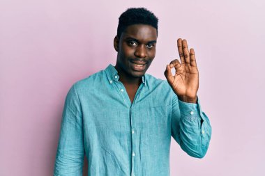 Yakışıklı, sıradan kıyafetler giyen, pozitif gülümseyen, el ve parmaklarıyla imza atan siyahi bir adam. başarılı ifade. 