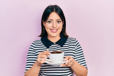 Bir fincan kahve içen genç İspanyol kadın mutlu ve serin bir gülümsemeyle gülümsüyor. diş gösterme. 