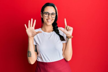 Genç İspanyol kadın profesyonel garson önlüğü giyip yedi numaralı parmaklarıyla işaret ederken kendine güvenen ve mutlu bir şekilde gülümsüyor.. 