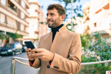 Zarif bir ceket giyen yakışıklı bir iş adamı akıllı telefon kullanarak dışarıda mutlu bir şekilde gülümsüyor.