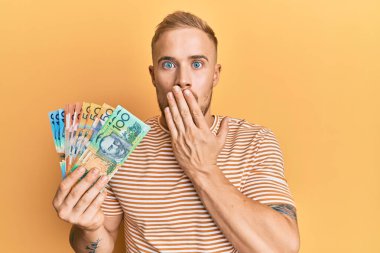Genç, beyaz bir adam Avustralya doları tutuyor. Ağzını eliyle kapatıyor. Şok olmuş ve hata yapmaktan korkuyor. şaşırmış ifade 