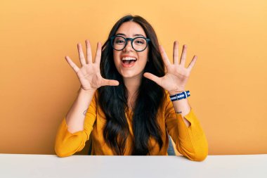 Güzel, esmer, gözlüklü ve gündelik giysileri olan genç bir kadın masanın üzerinde oturuyor ve parmaklarını on numarayı gösterirken kendinden emin ve mutlu bir şekilde gülümsüyor.. 