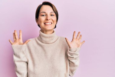 Kısa saçlı esmer kadın, günlük kışlık süveter giyiyor ve parmaklarıyla on numarayı gösteriyor. Kendinden emin ve mutlu bir şekilde gülümsüyor.. 