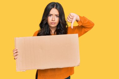 Güzel, esmer, elinde boş pankart tutan, kızgın yüzlü, olumsuz işaretli, baş parmakları aşağıda, reddedilme kavramından hoşlanmayan genç bir kadın. 