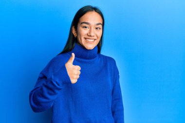 Sıradan bir kazak giyen genç Asyalı kadın elleriyle mutlu bir jest yapıyor. Kameraya bakıp başarıyı gösteren ifade. 