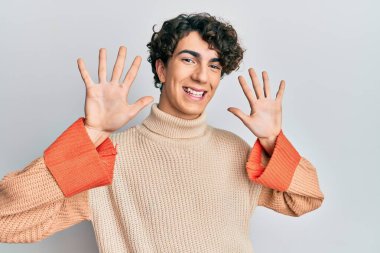 Hispanik genç adam, günlük kazak giyiyor ve parmaklarıyla on numarayı gösterirken kendinden emin ve mutlu gülümsüyor.. 