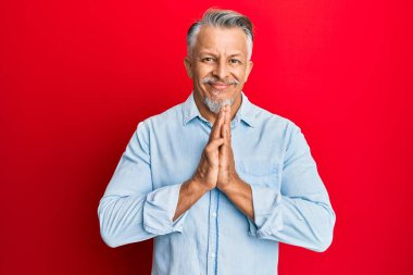 Orta yaşlı, gri saçlı, günlük kıyafetler giyen, elleriyle dua eden, affedilmek isteyen, kendinden emin, gülümseyen bir adam.. 