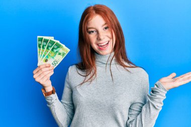 Genç kızıl saçlı kız 50 İsrail şekeri tutuyor başarıyı kutlayan banknotlar mutlu bir gülümsemeyle ve kazanan ifade kaldırılmış el ile. 