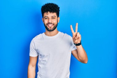 Sakallı, beyaz tişörtlü genç bir Arap. İki numaralı parmaklarıyla kendini beğenmiş ve mutlu bir şekilde iki numarayı gösteriyor.. 
