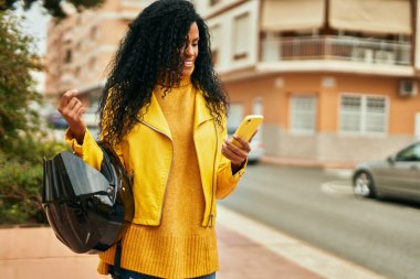 Orta yaşlı Afrikalı Amerikalı kadın motosiklet kaskı tutuyor ve şehirde akıllı telefon kullanıyor..