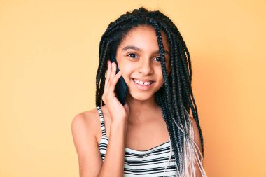Saçları örgülü genç Afro-Amerikalı kız akıllı telefondan konuşuyor. Olumlu ve mutlu görünüyor. Kendine güvenen bir gülümsemeyle dişlerini gösteriyor. 