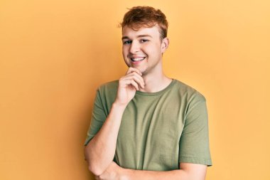 Gündelik yeşil tişört giyen genç beyaz bir adam kameraya kollarını kavuşturup çenesine dokunarak gülümsüyor. Olumlu düşünmek. 