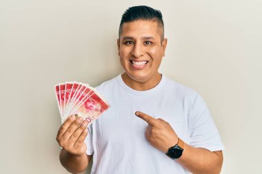Elinde 20 İsrail şekeri tutan genç latin adam banknotlar mutlu bir şekilde el ve parmağıyla işaret ediyor. 