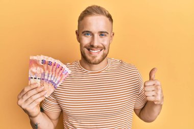 Güney Afrika 'nın 50 rand banknotunu elinde tutan genç beyaz adam mutlu ve olumlu gülümsüyor, başparmağıyla mükemmel ve onay işareti yapıyor. 