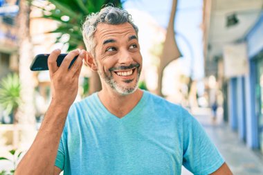 Orta yaşlı, kır saçlı, akıllı telefondan konuşan bir adam şehrin caddesinde yürüyor..