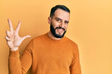 Sakallı genç adam, sıradan bir kazak giyiyor ve üç numaralı parmaklarını gösterip gülümsüyor. Kendinden emin ve mutlu.. 