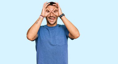 Dövmeleri olan, sıradan kıyafetler giyen, dürbün gibi hareket eden, dilini dışarı çıkaran, parmaklarından bakan yakışıklı bir adam. çılgın ifade. 