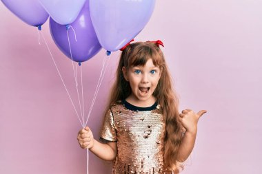 Kafkasyalı küçük kız elinde balonlarla baş parmağını kaldırmış ağzı açık gülümsüyor. 