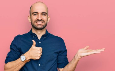 Latin asıllı genç adam, günlük kıyafetler giymiş, avuç içini gösteriyor ve baş parmaklarıyla iyi bir jest yapıyor, mutlu ve neşeli gülümsüyor. 