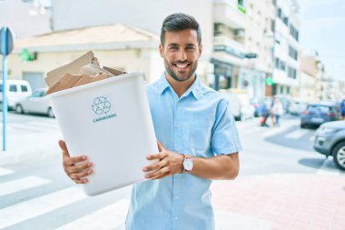 Mutlu geri dönüşümler gülümseyen İspanyol genç adam. Şehrin caddesinde bir kutu karton duruyor..